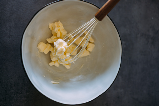 An overhead image of a whisk and butter in a bowl on a dark background.