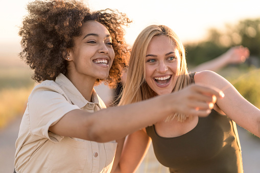 Close up of two young multiracial women walking on a road in the nature on sunset, pointing something in front of them