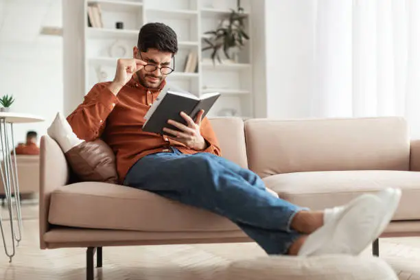Focused confused young Arabic man trying to read paper book, squinting to see more clearly, wearing glasses, having difficulties seeing text because of vision problems, sitting on couch in living room