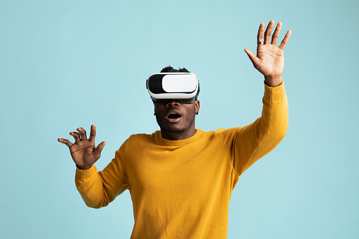Emotional young african american man in yellow sweater using VR glasses, touching something invisible, millennial black guy experiencing virtual reality adventure on blue studio background, copy space