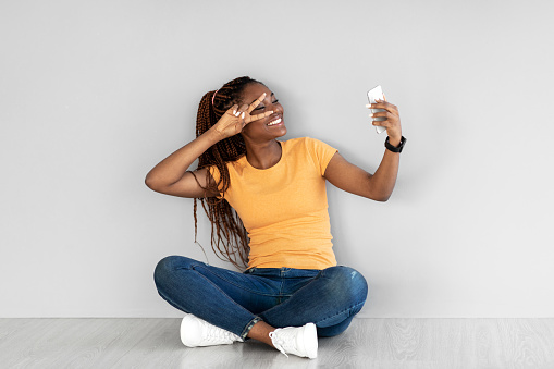 Attractive young African American woman with smartphone communicating online, showing peace gesture against grey studio wall, full length. Video chat, web meeting concept