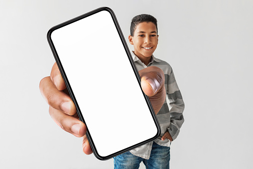 Happy African American Boy Holding Huge Smartphone With Empty Screen Advertising Mobile Application For Cellphone Standing Over Gray Studio Background. Kids, Technology And Gadgets. Mockup