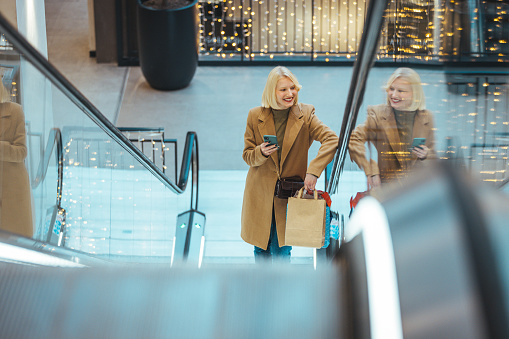 Beautiful Smiling Woman Using Mobile Phone while Standing on the Escalator. Cheerful young female typing text message on her smartphone and holding shopping bags in the shopping mall