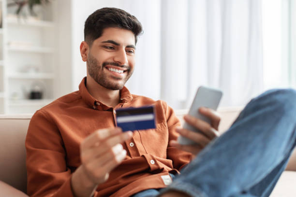 istock Smiling Arab guy using phone and credit card at home 1369129876