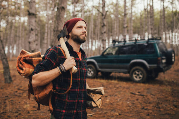 istock Millennial man hipster with beard wearing backpack, holding axe and firewood in forest with car on background. 1369130048