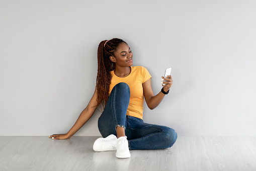 Full length of young African American lady sitting with cellphone, browsing internet, chatting online against grey studio wall. Millennial black woman using mobile phone, checking social media
