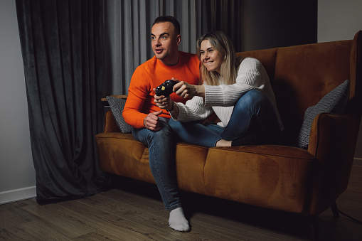 young cheerful family playing games with joystick on comfortable brown sofa. Looking at TV and rejoice in victory. Screaming with joy in gray tone room. Wear colorful sweeter, blue jeans.
