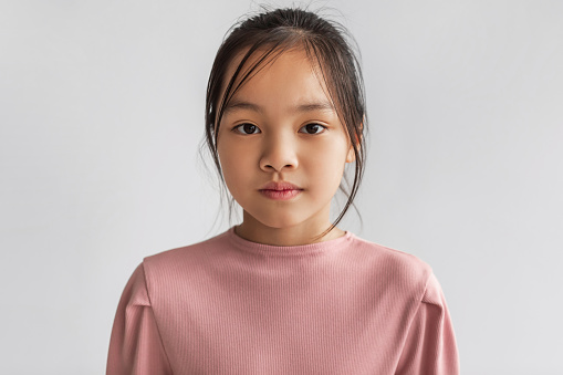 Portrait Of Serious Asian Girl Looking At Camera Posing On Gray Background In Studio, Wearing Pastel Pink Longsleeve. Children Fashion And Clothing Concept. Front View Shot