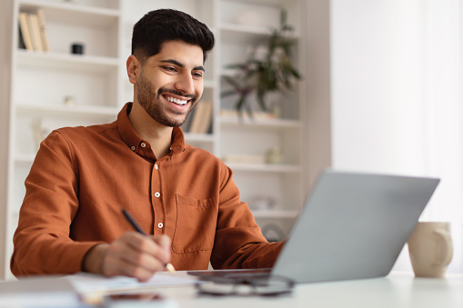 Portrait of young smiling Arab man using laptop sitting at desk, writing in notebook. Cheerful guy browsing internet, watching webinar studying online, looking at pc screen, free copy space