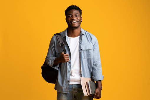 Positive millennial black man student in casual posing with books and notepads on yellow studio background, young african american guy enjoying studying at university or college, copy space