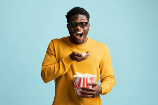 Excited black guy eating popcorn and smiling, using 3d glasses Excited young black guy in yellow eating popcorn and smiling, using 3d glasses, watching newest movies at cinema, posing over blue studio background, copy space. Entertainment concept 3 d glasses stock pictures, royalty-free photos & images