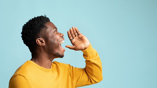 Happy african american guy posing over blue studio background, holding hand to mouth and saying something out loud, panorama with copy space. Handsome young black man making announcement, side view