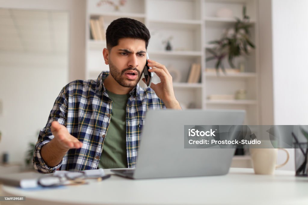 Confused Arab man talking on mobile phone using laptop Confused Discontented Arab Man Talking On Phone Using Laptop Having Problem With Computer Or Internet Connection At Home. Displeased Male Customer Calling To Hotline Service And Complaining Frustration Stock Photo