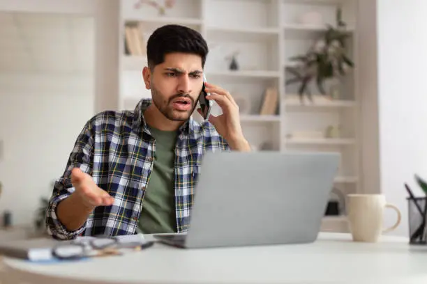 Confused Discontented Arab Man Talking On Phone Using Laptop Having Problem With Computer Or Internet Connection At Home. Displeased Male Customer Calling To Hotline Service And Complaining