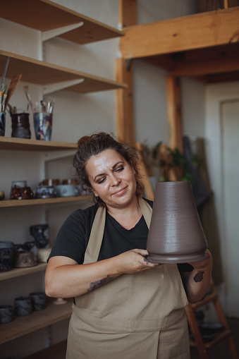 A middle-aged plus size woman in a pottery apron creates a clay vase on a potter's wheel in a pottery workshop