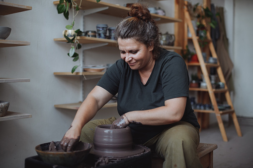 A middle-aged plus size woman creates a clay vase on a potter's wheel in a pottery workshop
