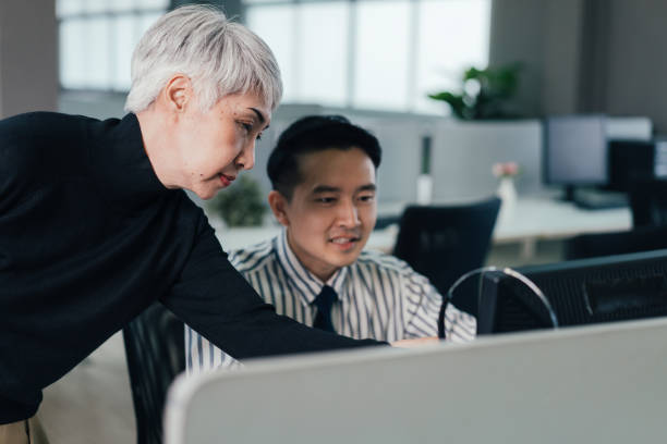 istock Asian senior manager checking and discussing project on computer with male employee in the office. corporate working at desk overtime together 1369141477