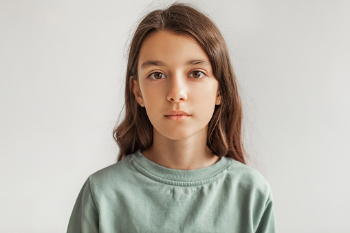 Portrait Of Serious Preteen Girl Standing Looking At Camera Over Gray Studio Background. Calm Kid Posing Wearing Casual Clothes, Headshot. Children Fashion Concept. Front View