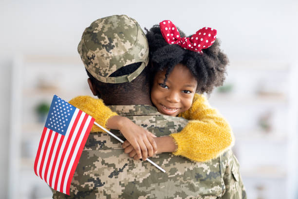 piccola ragazza nera carina che tiene la bandiera americana e abbraccia il papà del soldato - homecoming foto e immagini stock