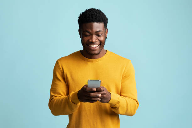 Joyful african american guy holding smartphone and smiling Joyful african american millennial guy holding modern smartphone, looking at gadget screen and smiling on blue studio background, black man gambling or gaming online, texting girlfriend, copy space people using mobile phone stock pictures, royalty-free photos & images