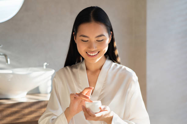 Smiling young asian lady holding jar with beauty product Smiling attractive young asian lady in white bathrobe holding jar with beauty product, enjoying morning routine for beautiful face skin, standing in modern bathroom, sun flare, copy space woman moisturizer stock pictures, royalty-free photos & images