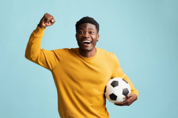 emotivo chico negro con balón de fútbol posando sobre azul - fan fotografías e imágenes de stock