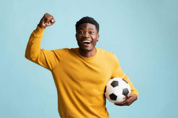 un gars noir émotif avec un ballon de football posant sur du bleu - soccer player soccer men smiling photos et images de collection