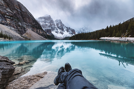 Legs of traveler relaxing on Moraine Lake with rocky mountains in gloomy day at Banff national park, Alberta, Canada