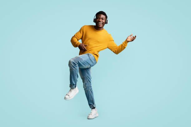 Carefree young black guy with wireless headset dancing Carefree young black guy in casual with wireless headset dancing and listening to music, imitating guitar playing, enjoying newest stereo headphones, blue background, full length, copy space dancer stock pictures, royalty-free photos & images
