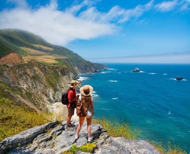 Couple on hiking trip resting on top of the mountain looking at ocean view. Friends relaxing on hiking trip by the Pacific ocean. People enjoying beautiful coastal scenery. People looking at beautiful ocean view. Pacific Ocean, Big Sur, California, USA american tourism stock pictures, royalty-free photos & images