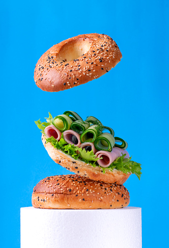 Flying bagel sandwich with ham and cucumber rolls and lettuce against the blue background. Levitating food