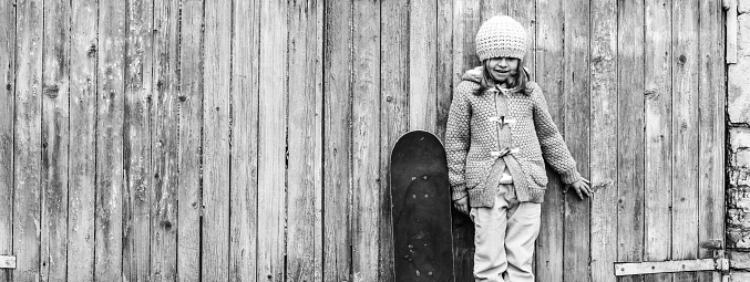 Horizontal banner or header with cute child girl in winter wool clothes with skateboard while standing against grunge wooden background - Concept of activity and playful childhood - Copy space