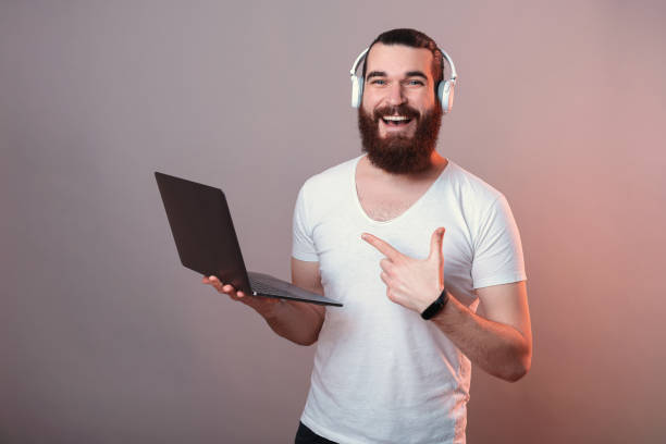 szeroko uśmiechnięty mężczyzna wskazuje na laptopa w słuchawkach. - working man audio zdjęcia i obrazy z banku zdjęć