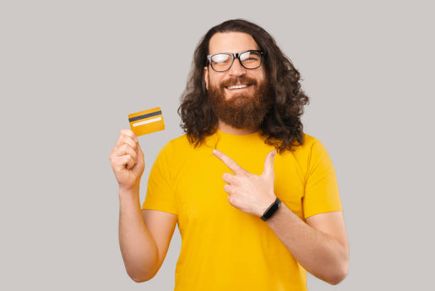 istock Joyful bearded man wearing glasses and pointing at yellow debit card over grey backdrop 1369166199