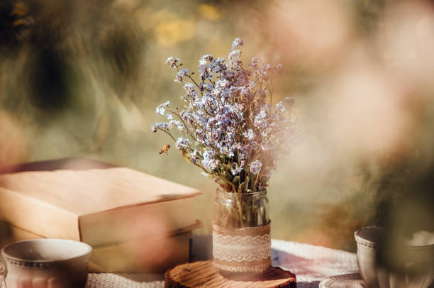 Dream like view through apple blossoms to table, set with tea cups and books. Bouquet of wildflowers Myosotis arvensis forget me nots in handmade jar vase.