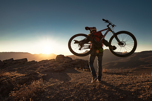 Sporty man cyclist climbs uphill with mountain bike at sunset
