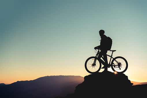 Cyclist's silhouette standing on big rock against sunset. Mountain bike concept