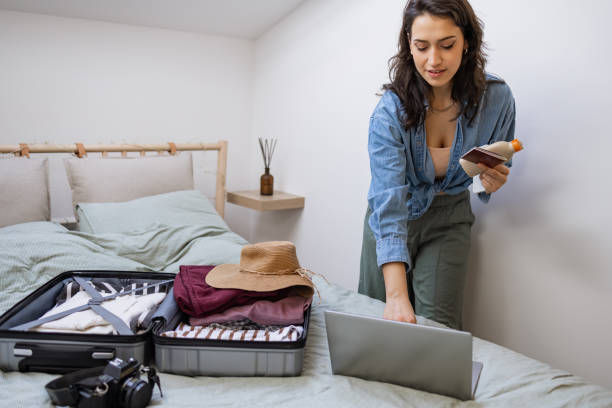 istock Young woman working on the laptop while packing her clothes for a trip 1369172950