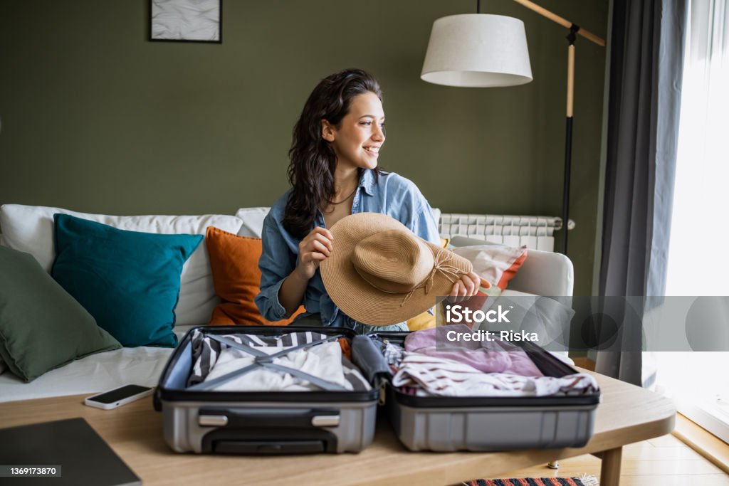 Portrait of a woman preparing for a trip Young woman sitting on the couch and packing suitcase. She is holding a straw hat in her hands Travel Stock Photo