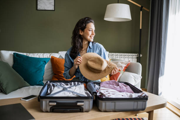 Portrait of a woman preparing for a trip Young woman sitting on the couch and packing suitcase. She is holding a straw hat in her hands packing stock pictures, royalty-free photos & images