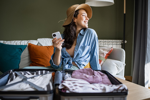 Young woman sitting on the couch and packing suitcase. She is holding a smart phone in her hand and wearing a straw hat.