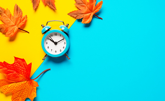 Clock on two tone color yellow and light blue background decorated with colorful fallen leaves. Autumn time concept. Copy space, flat lay