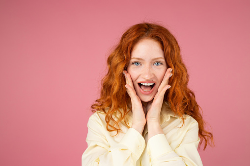 Excited redhead beautiful girl with blue eyes and opened mouth keeping her palms on cheeks posing over a pink background. High quality photo