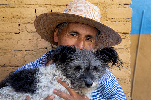 A cheerful looking gardener, wearing a straw hat, holding his little dog in front of a yellow wall