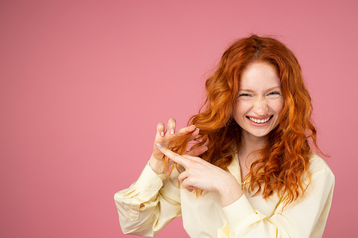Joyful redhead smiling lady in yellow dress holding her hair in hand and showing cut sign with two fingers over pink background and copy space for advertisement, hairstyle concept. High quality photo