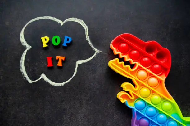 Photo of Pop it dinosaur toy rainbow colors on a black background with multicolored letters and the inscription - Pop it in a speech bubble