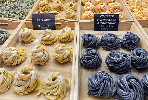 A variety of handmade spaghetti is sold by weight on food court. In Russian are written varieties of pasta: fettuccine, pasta with cuttlefish ink, and price.