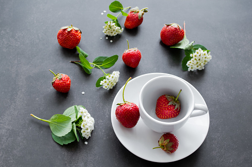 White spirea flowers and bright red strawberries in a white cup and scattered around on a black background. Summer mood background.