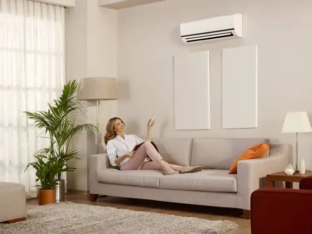 Young woman sitting and using remote control to control air conditioner in the house