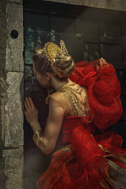 Beautiful young blonde woman wearing an elegant red and gold princess gown and golden jewels, looking through the key hole of a dungeon door The model is wearing traditional tembleque from Panama woman spying through a keyhole stock pictures, royalty-free photos & images